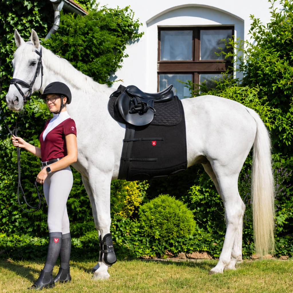 Woman in equestrian attire standing next to a white horse in a garden setting.